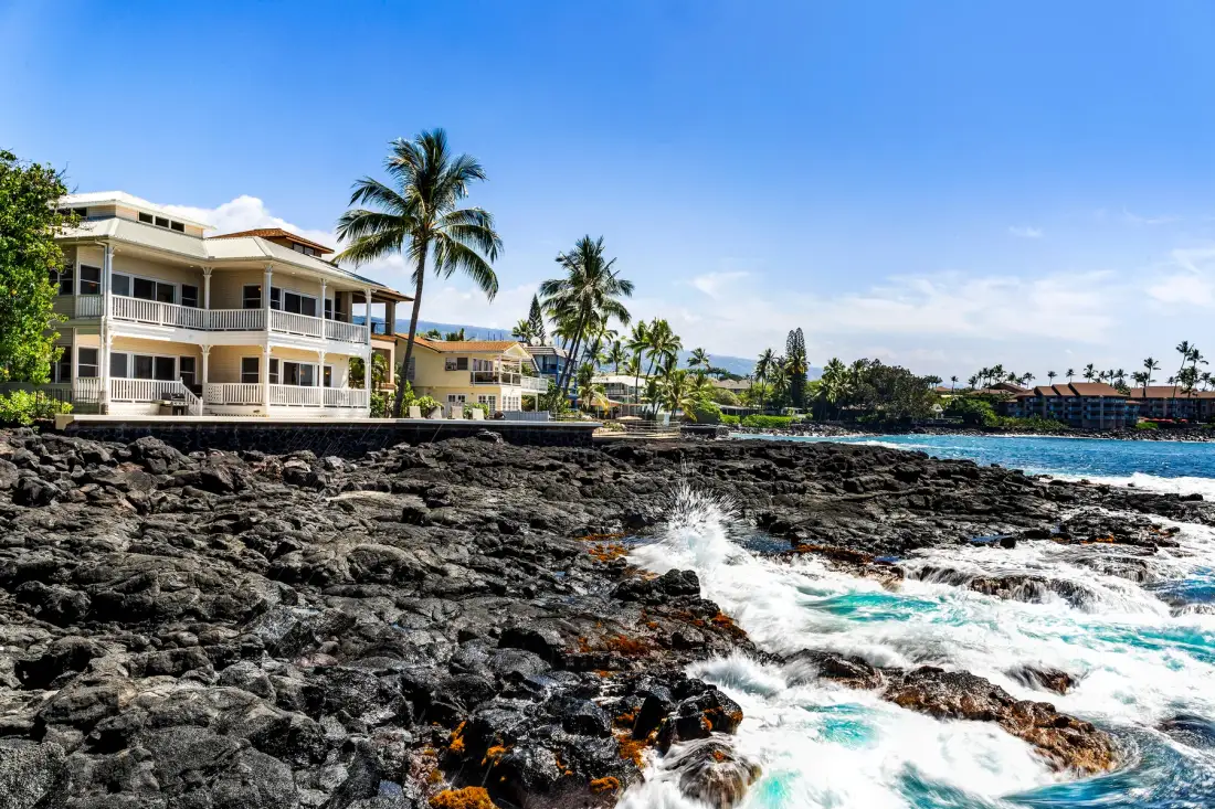 Las olas rompen en una costa rocosa junto a palmeras y edificios frente al mar.