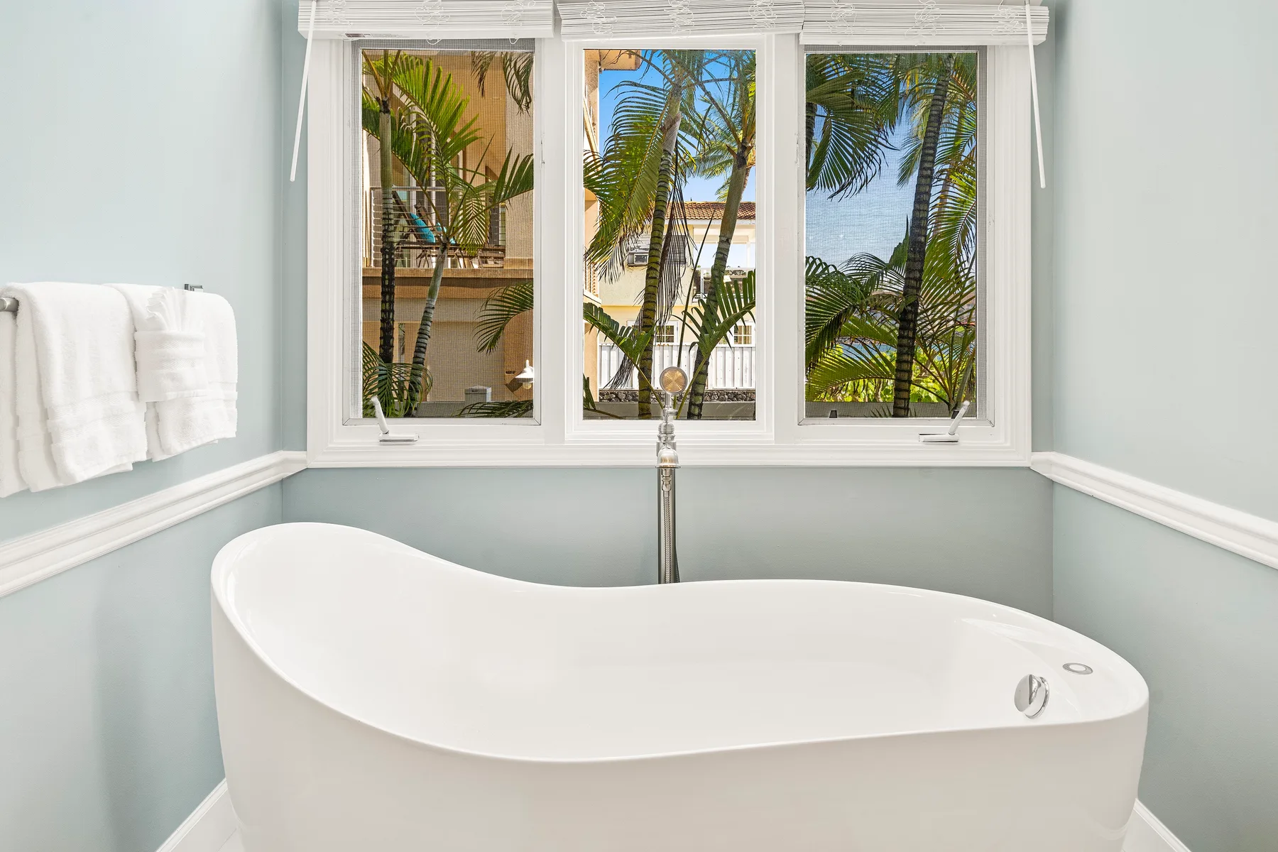 Una bañera exenta está bajo ventanas con vistas a palmeras.
