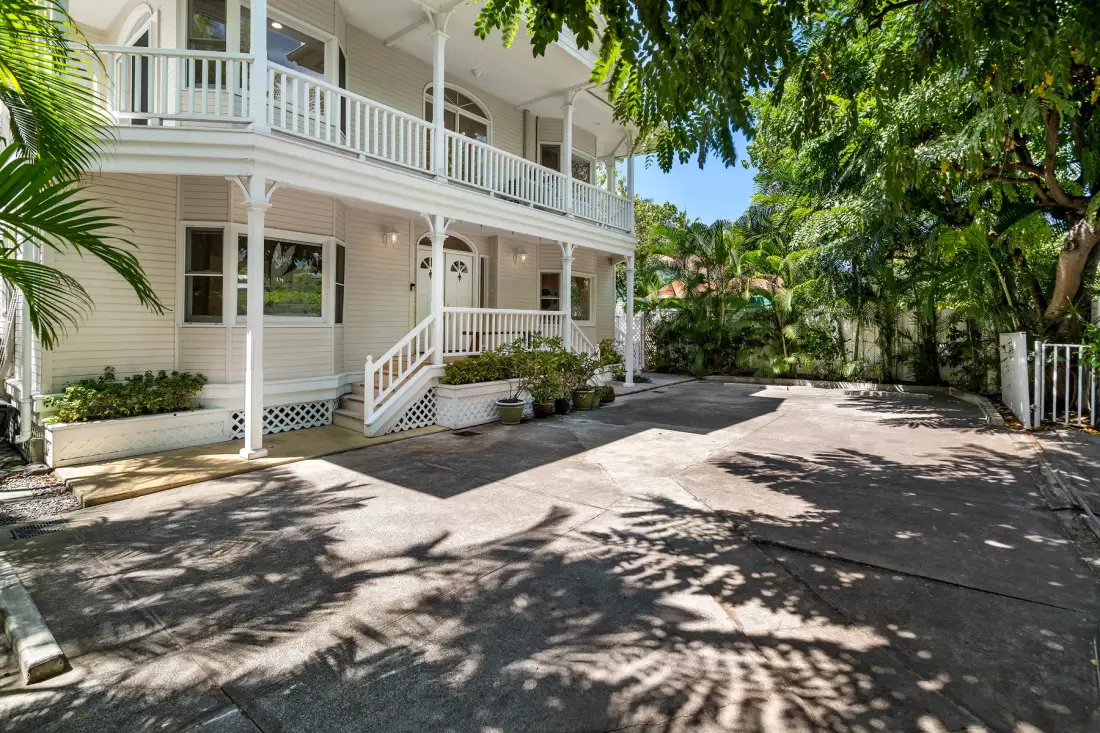 Una casa blanca de dos pisos con balcones da a una entrada pavimentada y sombreada por árboles.