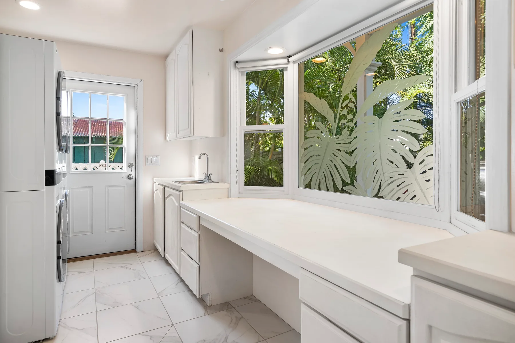 Una cocina luminosa con encimeras blancas y ventanas con vistas a vegetación frondosa.