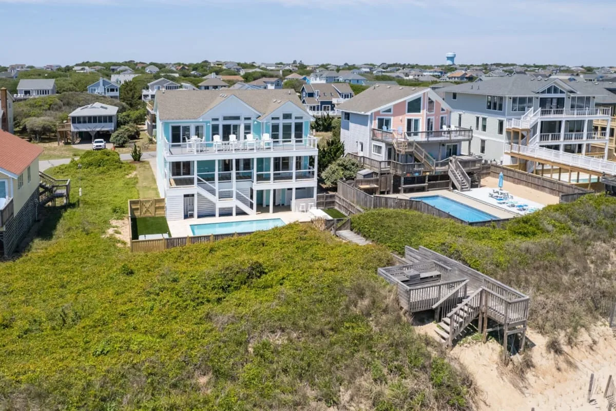 An aerial view of coastal houses with decks and a swimming pool beside grassy dunes.