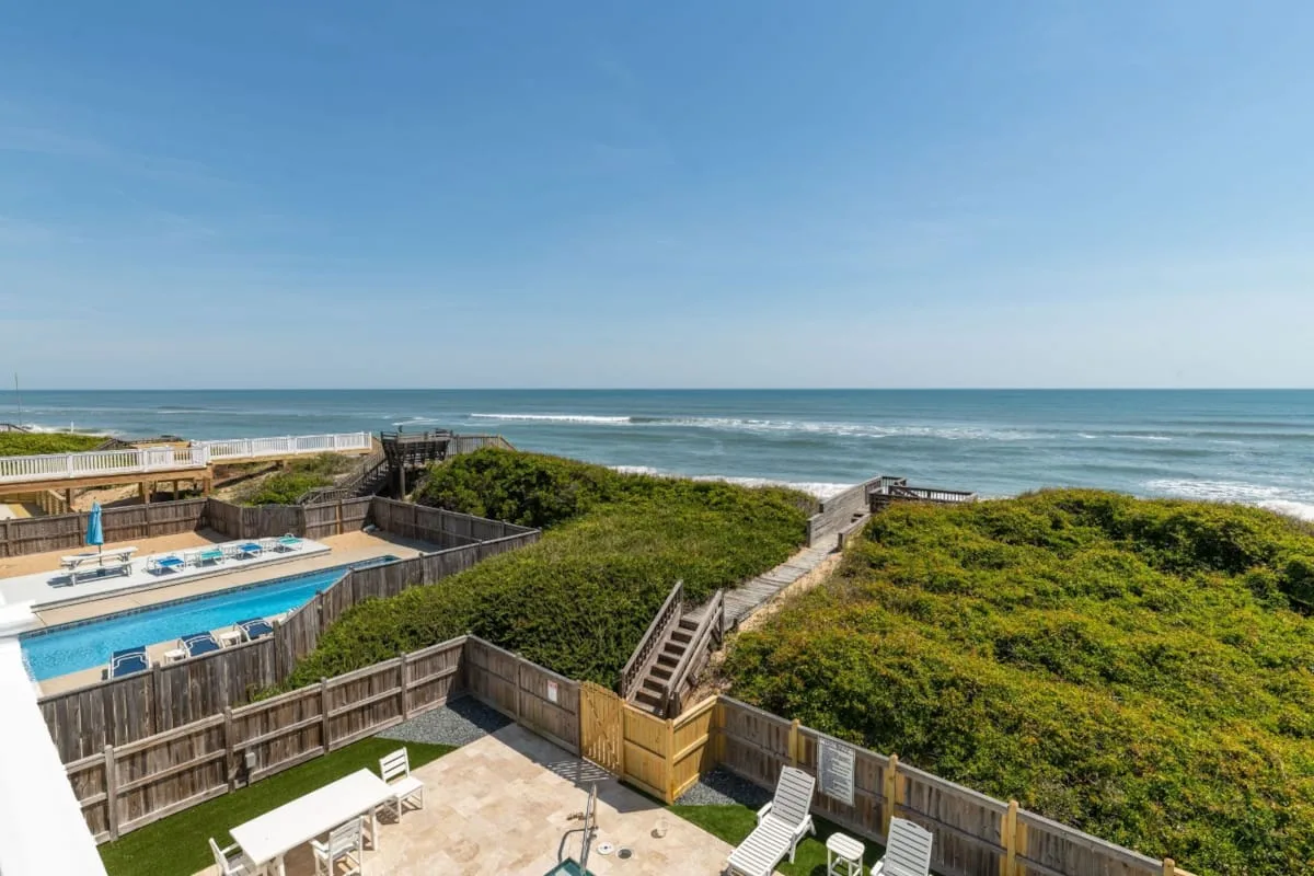 A fenced outdoor pool and patio sit beside green dunes overlooking the ocean.