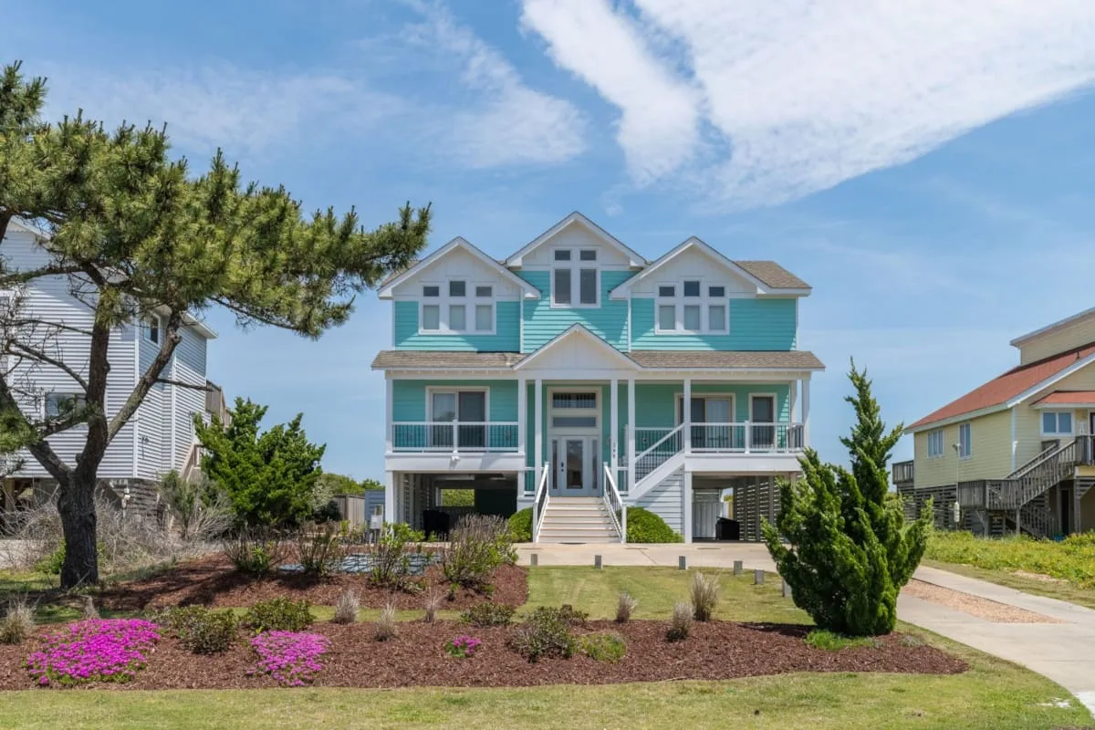 A large turquoise house with white trim stands beside a landscaped yard.