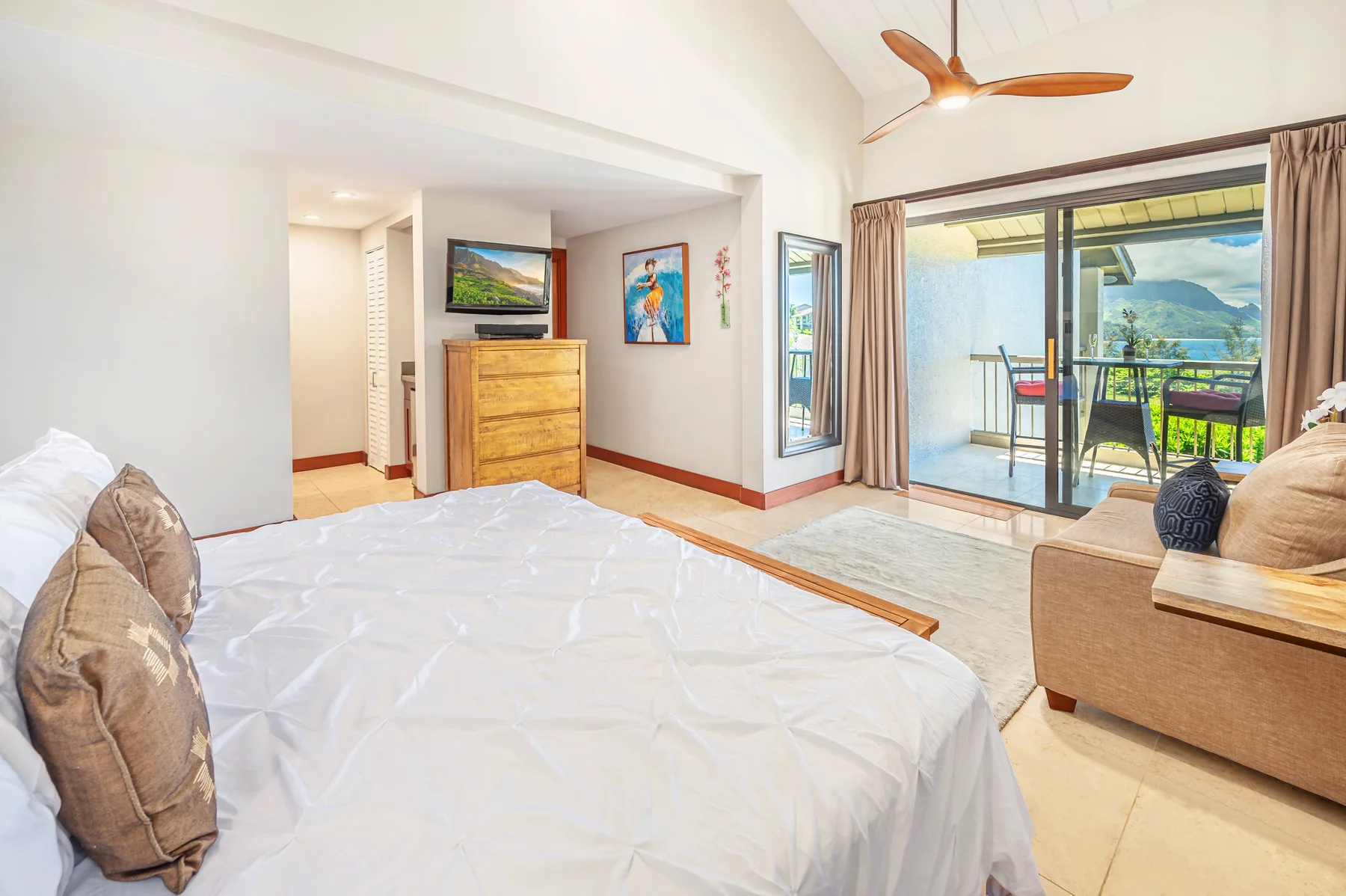 A bedroom with a bed, a wooden dresser, and sliding glass doors opening to a balcony.