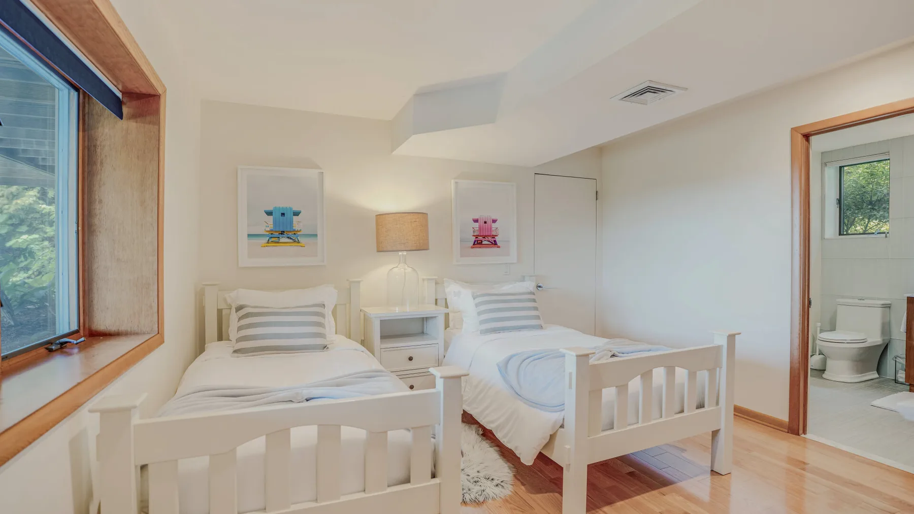 A bright bedroom with two beds, bedside tables, and an open doorway to a bathroom.