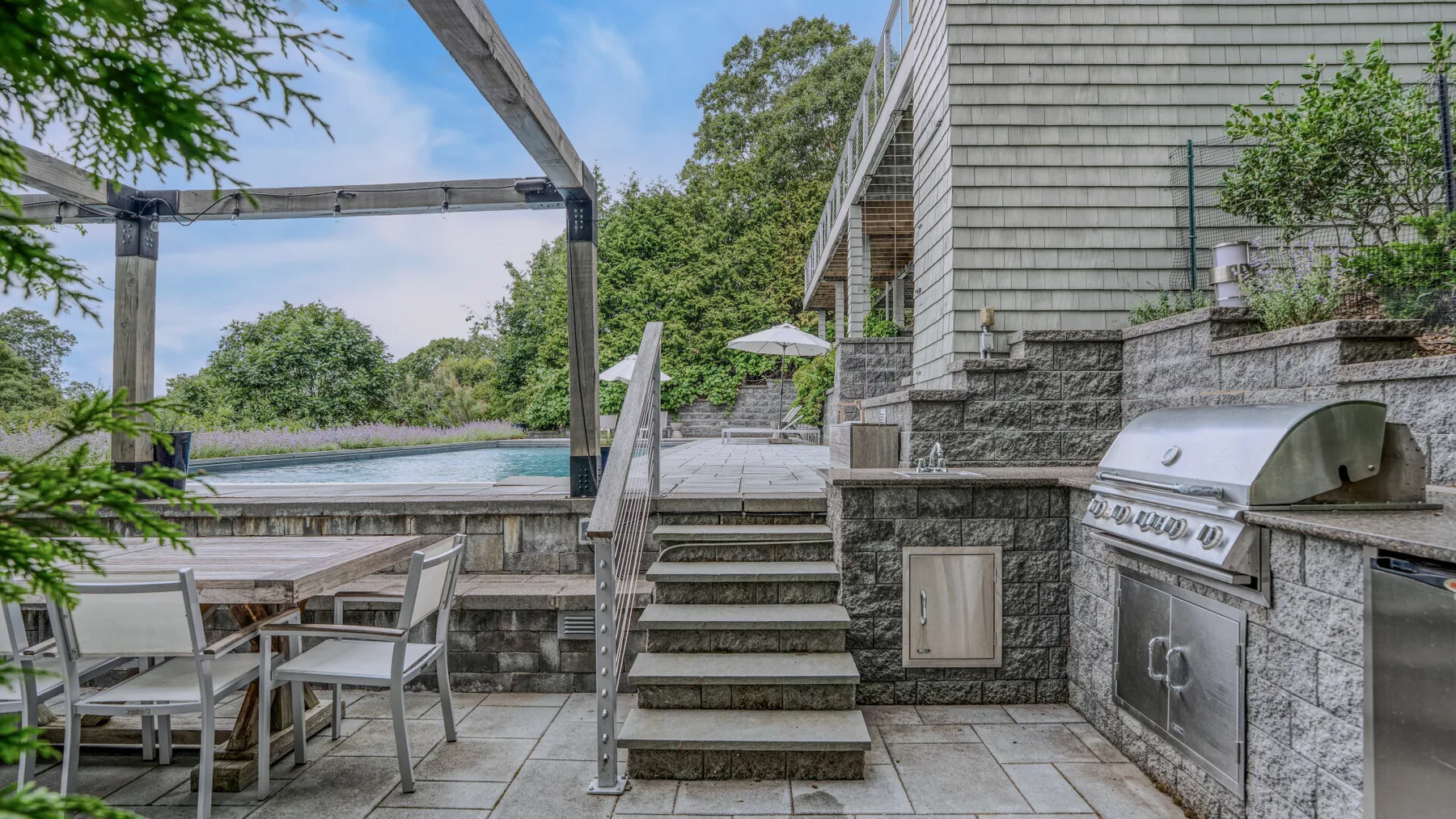 A stone patio with outdoor seating, steps, and a built-in grill overlooks a pool.