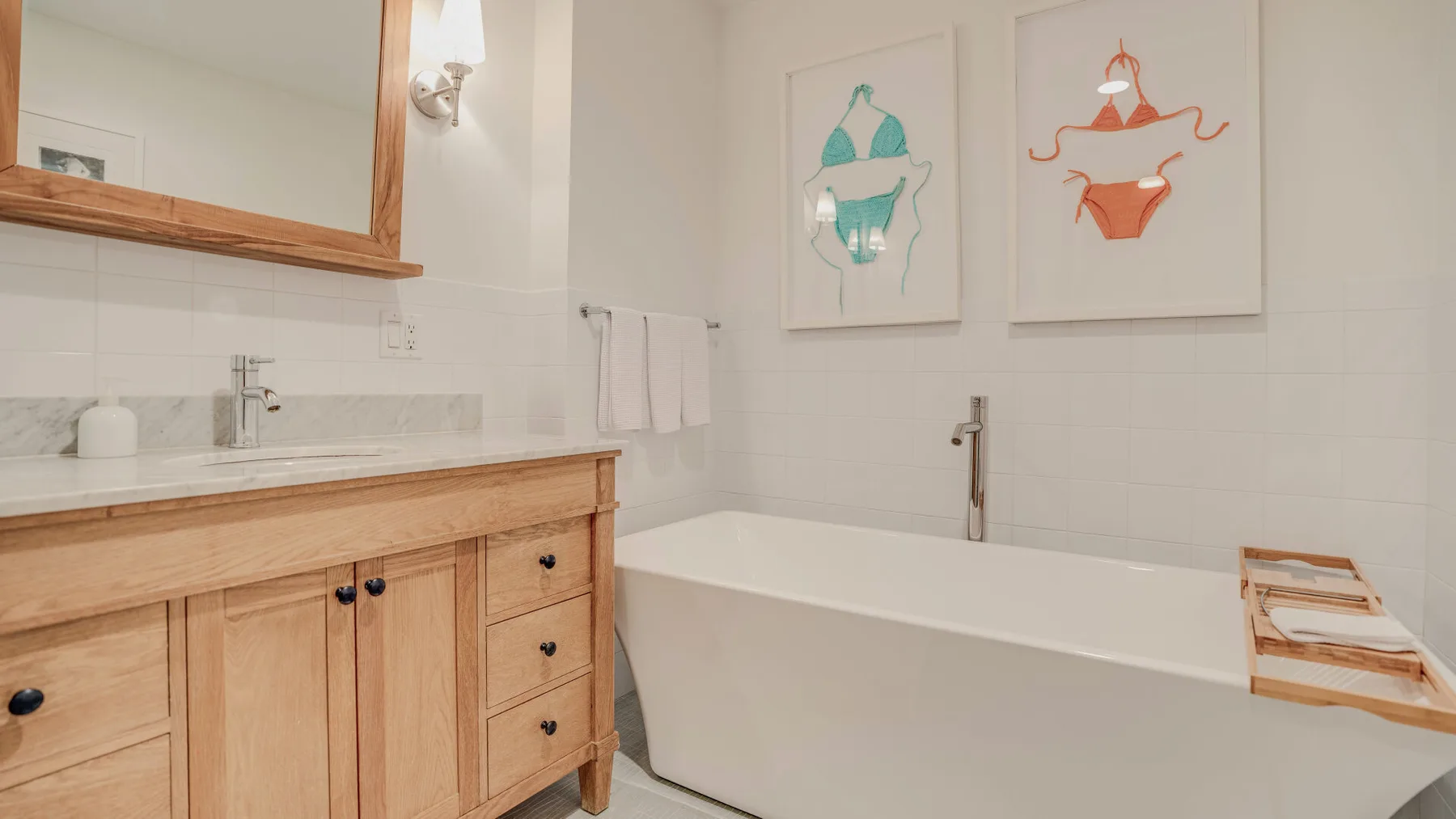 A bright bathroom with a wooden vanity, bathtub, and framed swimsuit prints.