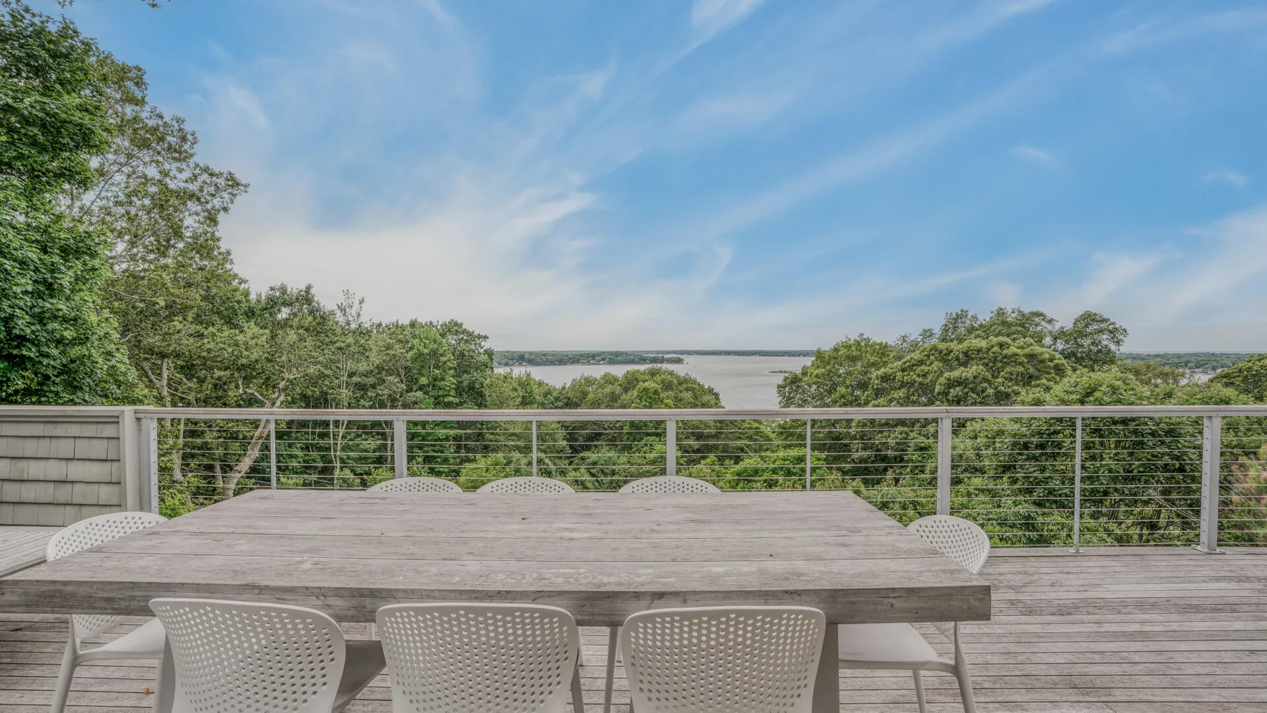 A wooden deck with an outdoor dining table overlooks green trees and a broad water view.