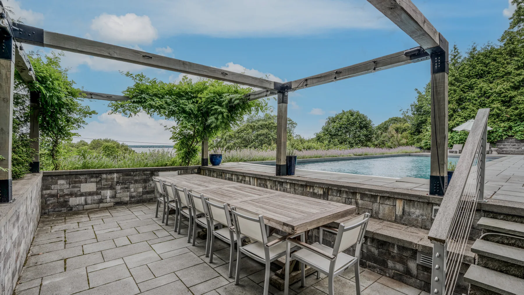 Una zona de comedor al aire libre con una mesa larga da a una piscina y vegetación.