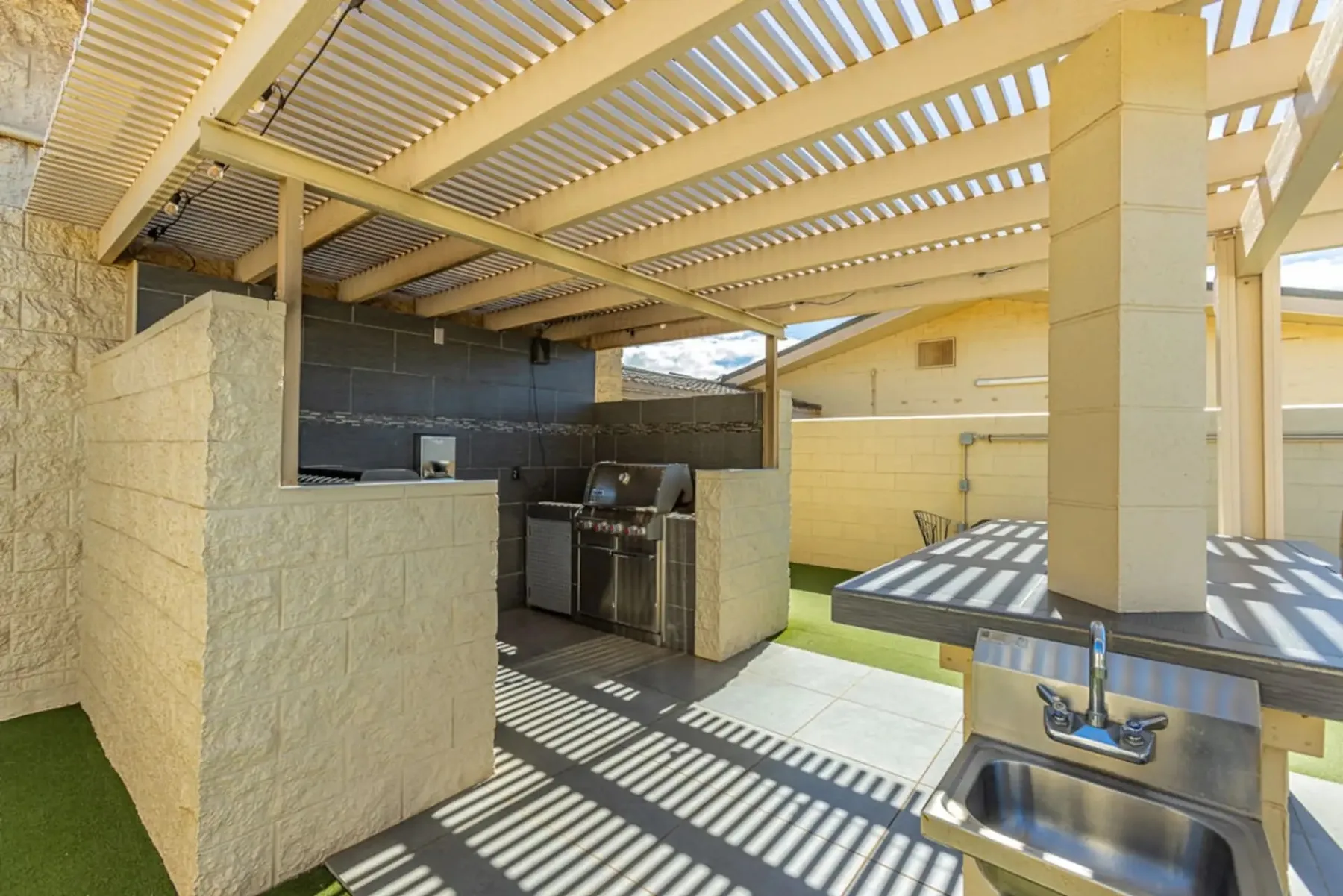 Una zona de cocina al aire libre bajo una pérgola de listones, con parrilla y fregadero.