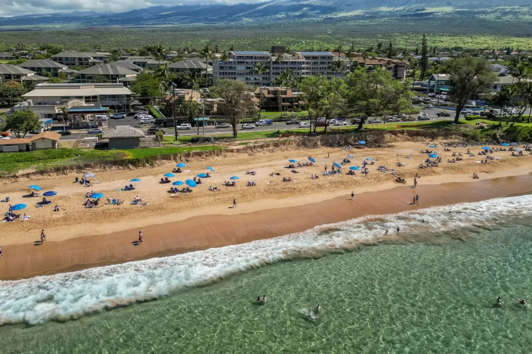 Vista aérea de una playa de arena con sombrillas azules y olas junto a la orilla.