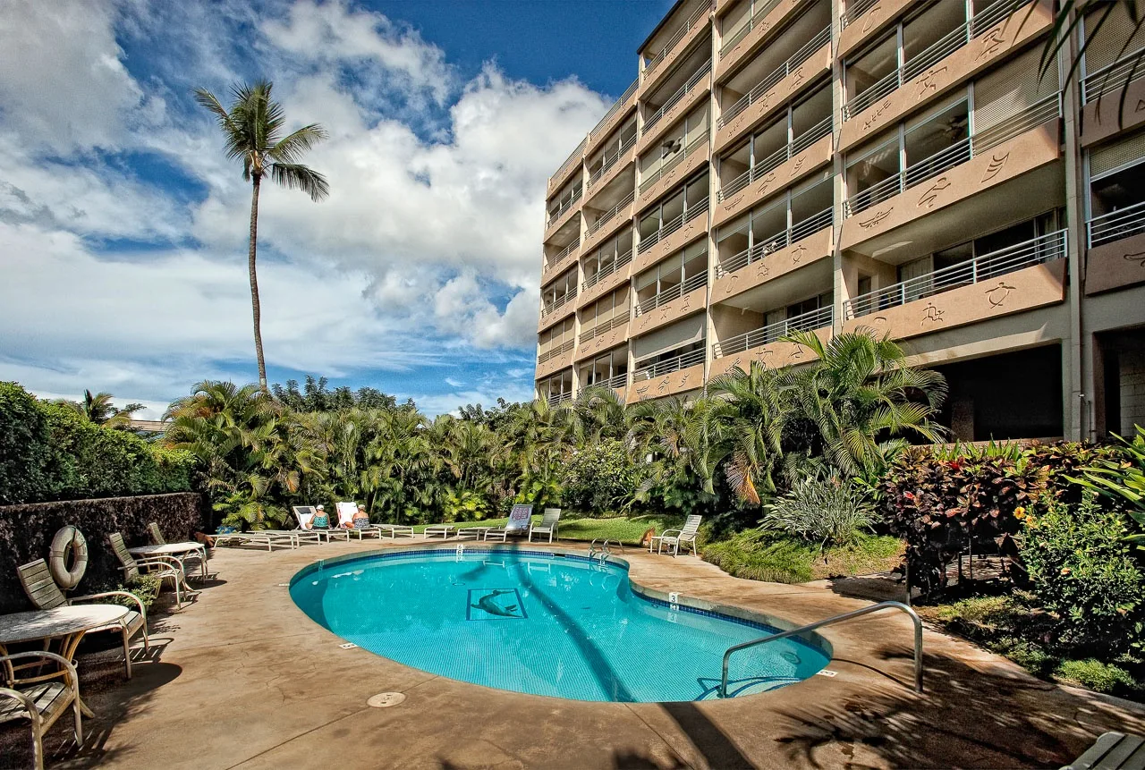 Una piscina al aire libre está junto a un edificio de varios pisos y vegetación tropical.