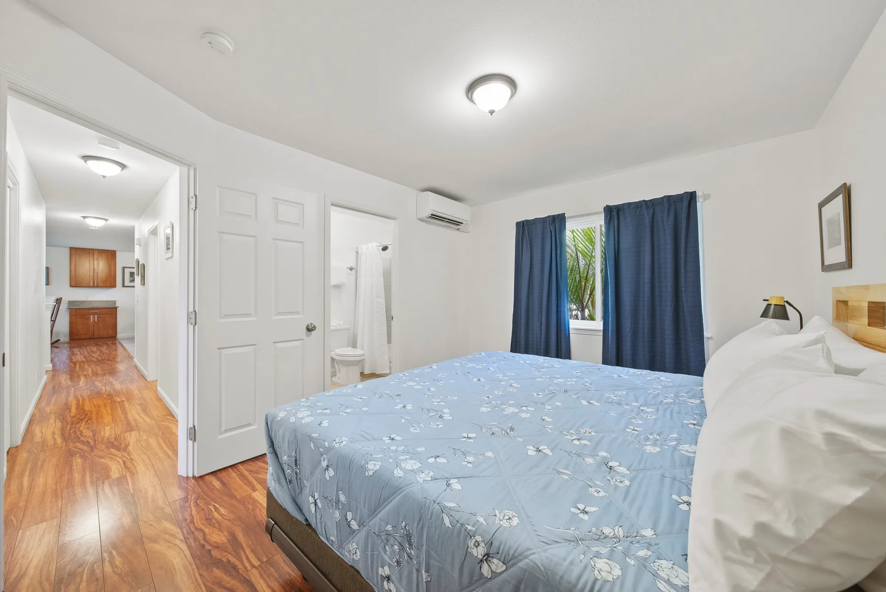 A bedroom with a blue floral bedspread, dark curtains, and a view into the hallway.
