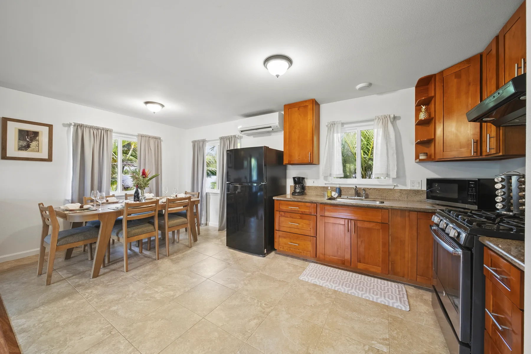 Cocina y comedor con armarios de madera, una mesa y ventanas grandes.