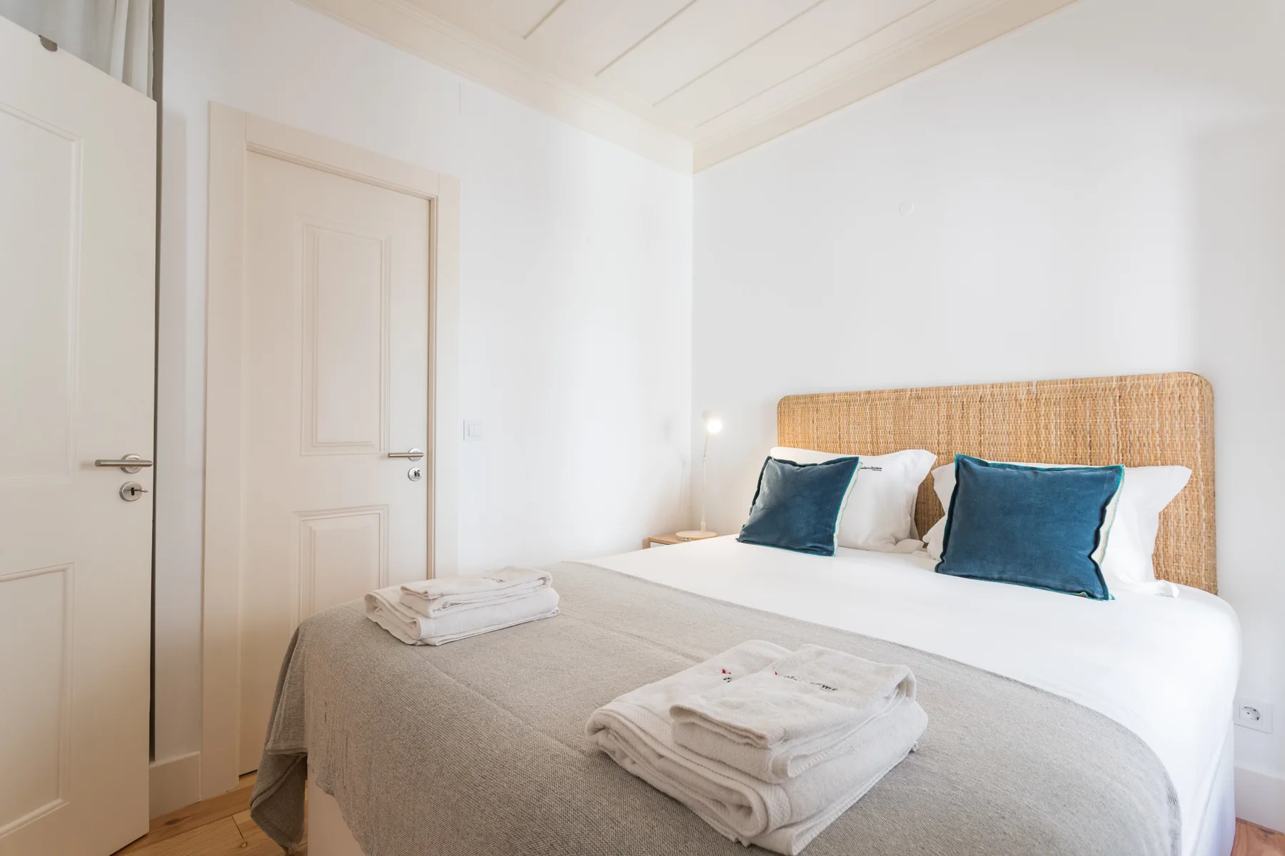 Un dormitorio luminoso con una cama, cabecero tejido y toallas dobladas.