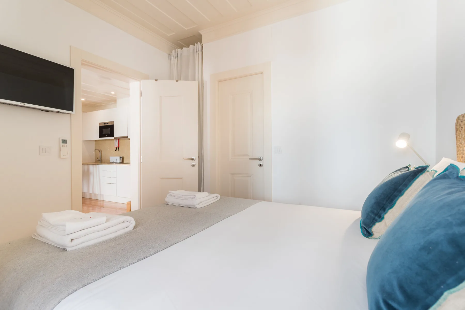 Un dormitorio luminoso con una cama, toallas dobladas y una puerta abierta a una pequeña cocina.
