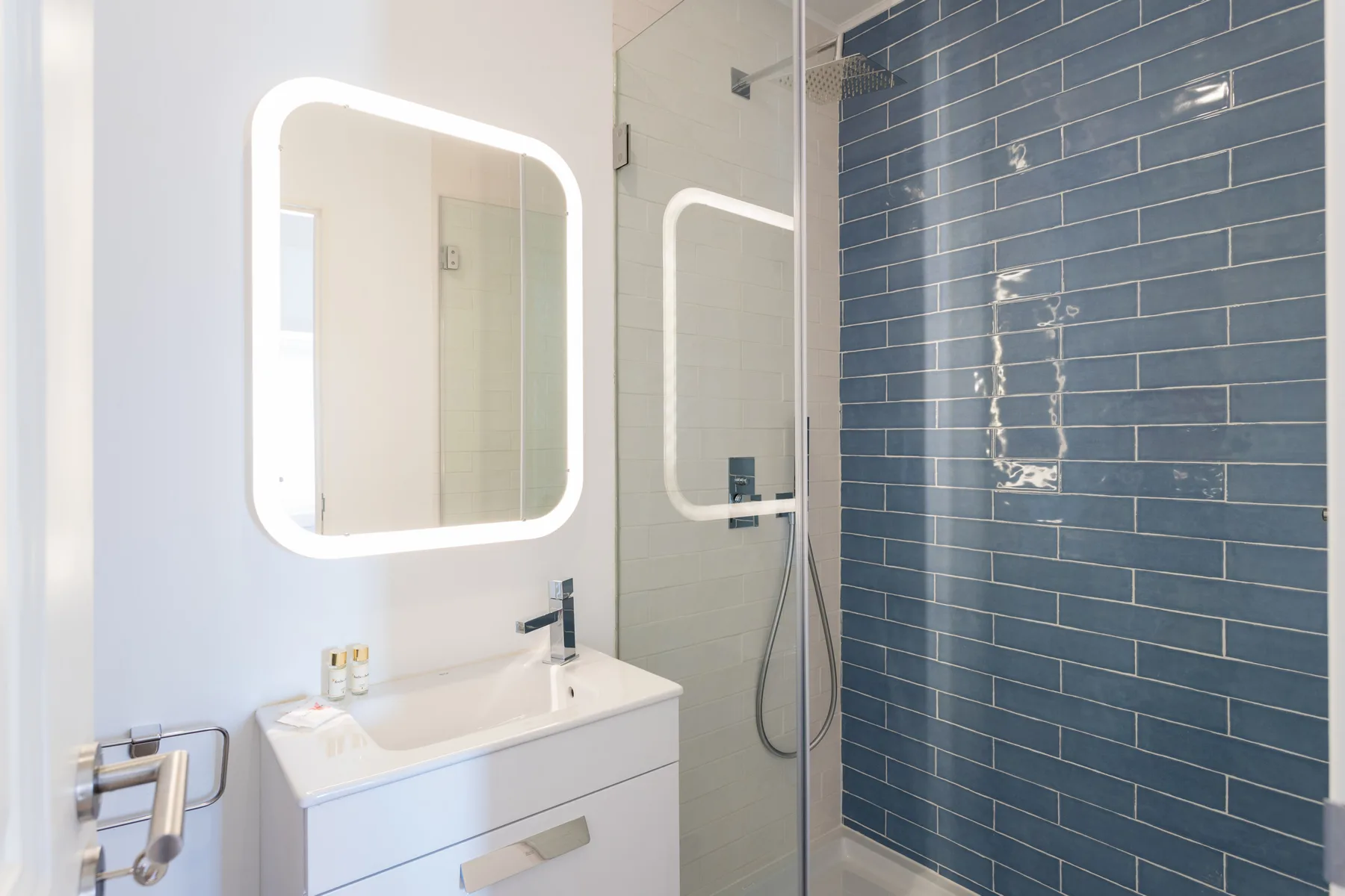 Un baño con lavabo blanco, espejo iluminado y ducha con azulejos azules.