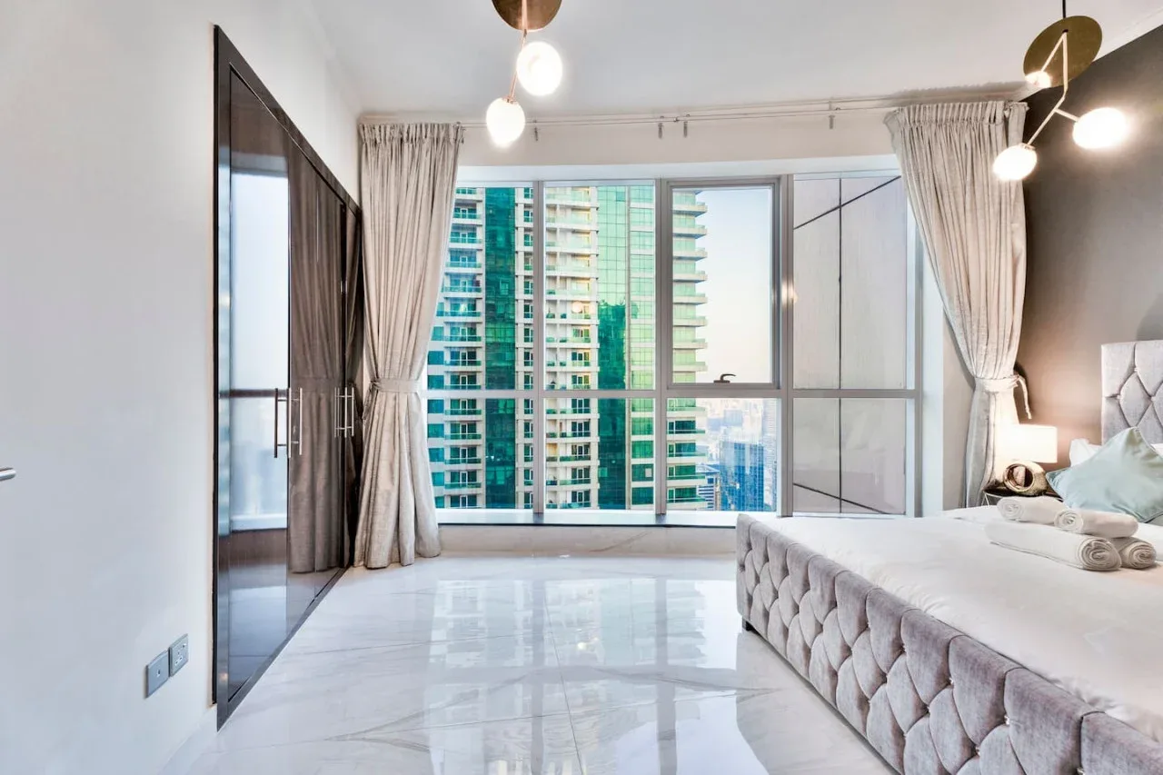 A bright bedroom with a large window overlooking nearby high-rise buildings.