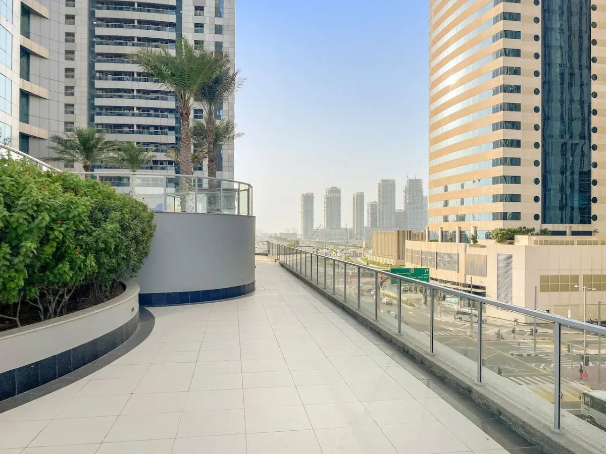 A tiled outdoor walkway runs between high-rise buildings, with glass railings and greenery.