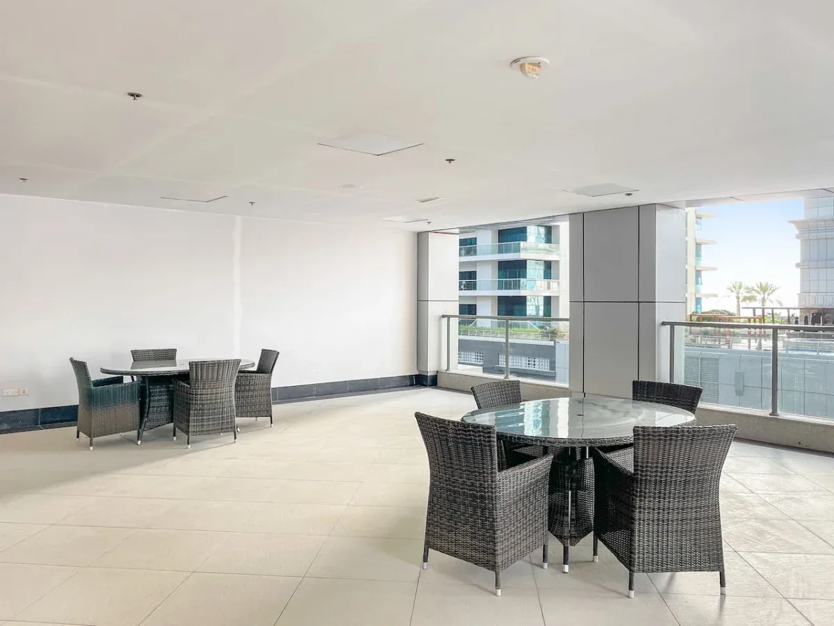 A bright covered terrace with seating and glass railings overlooking nearby buildings.