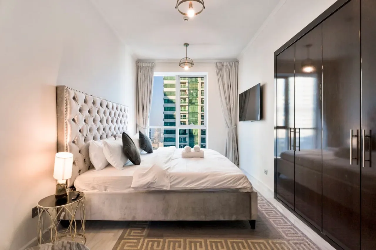 A bright bedroom features a large bed beside a window and dark wardrobes.