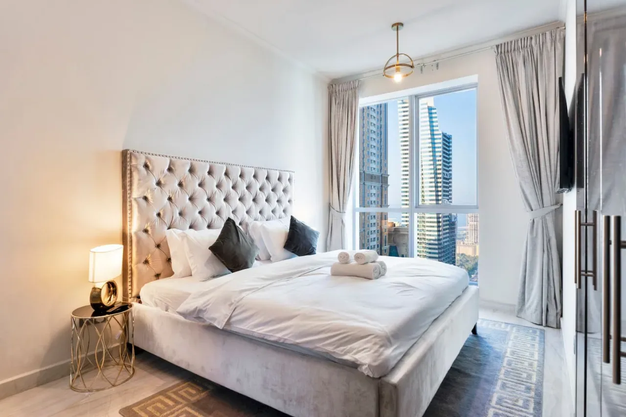 A bright bedroom with a large bed beside a window overlooking nearby buildings.