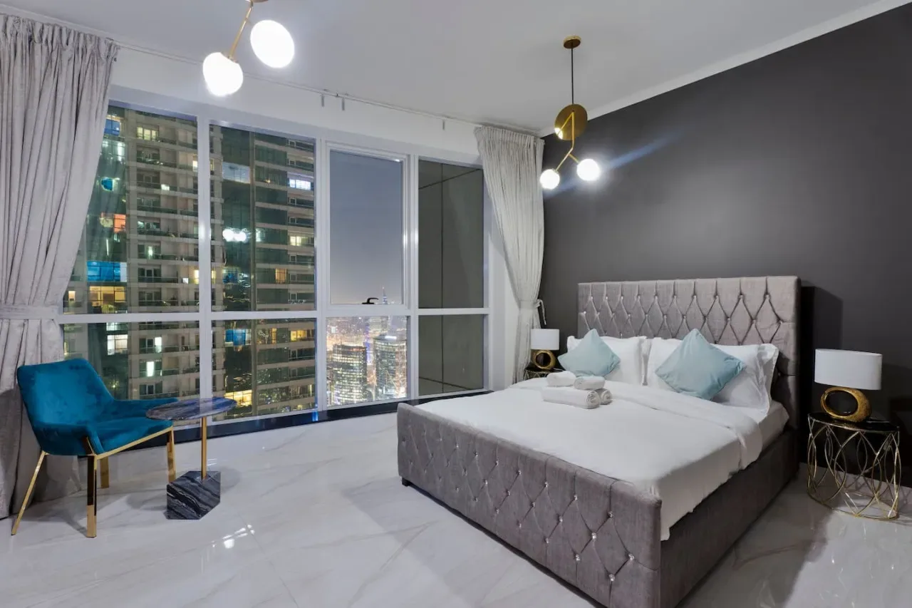 A bedroom with a large bed beside floor-to-ceiling windows overlooking city buildings.