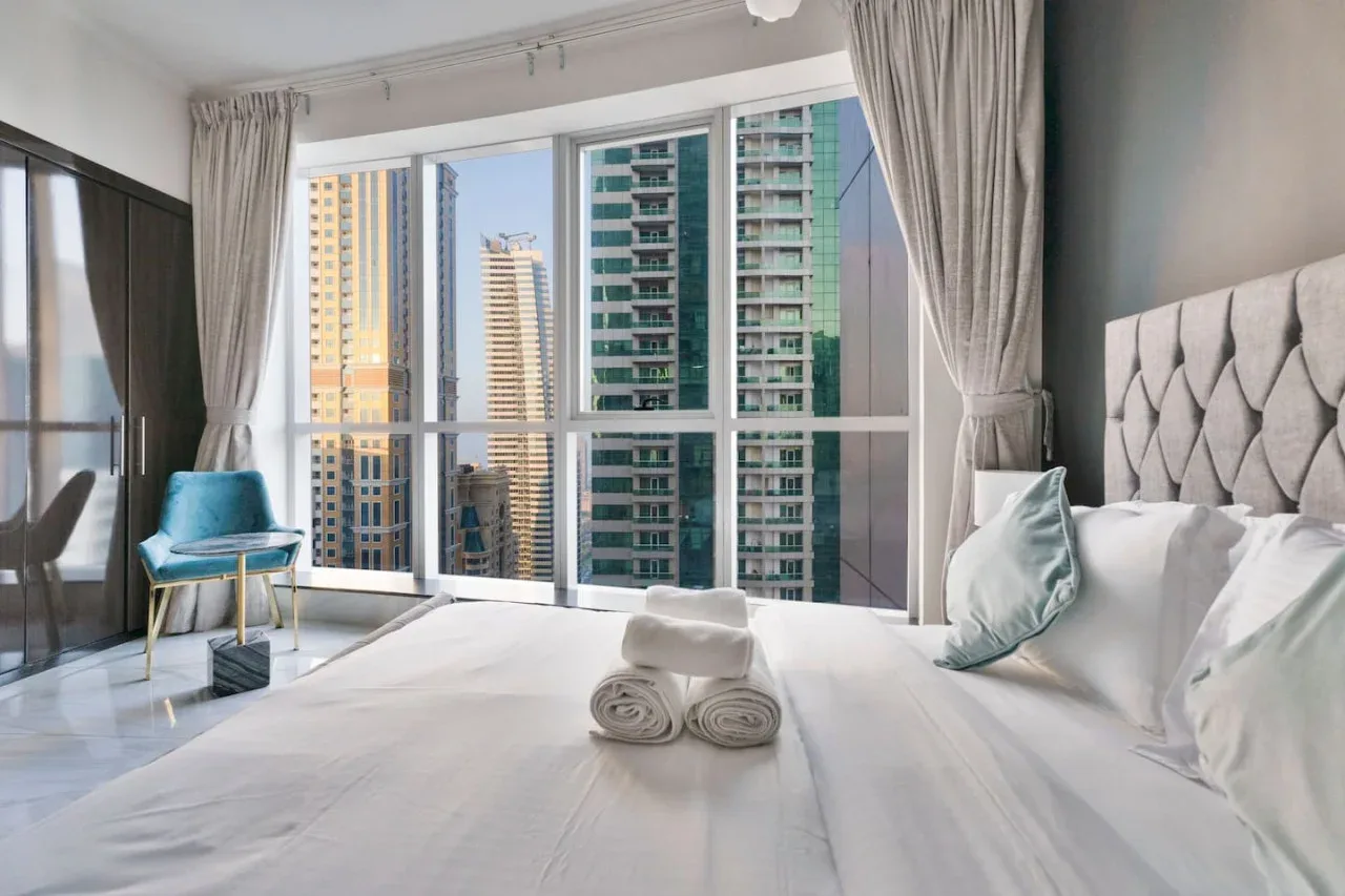 A bright bedroom with a neatly made bed and floor-to-ceiling windows overlooking nearby buildings.