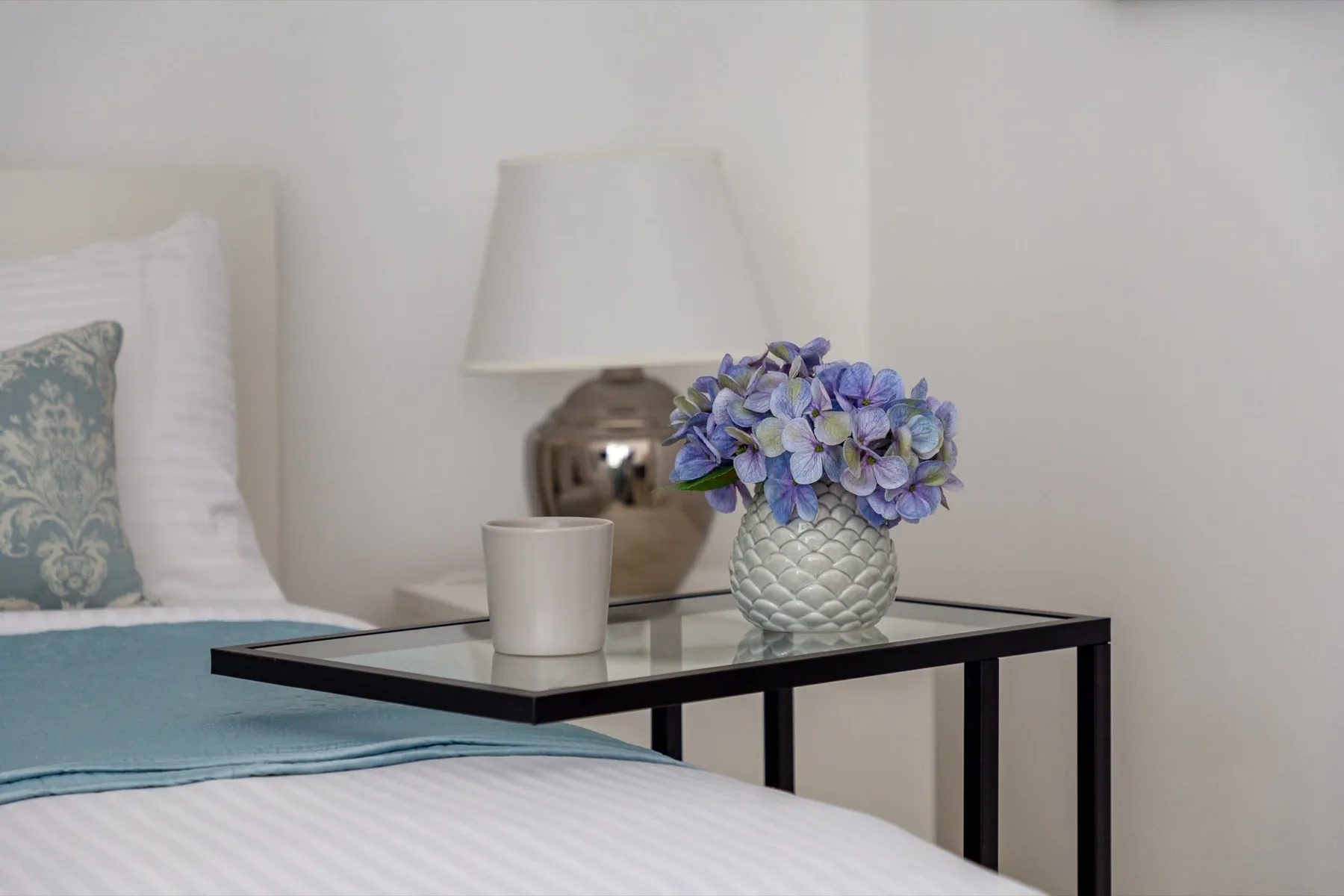 Un dormitorio con una mesa junto a la cama que sostiene una lámpara y un jarrón con flores.