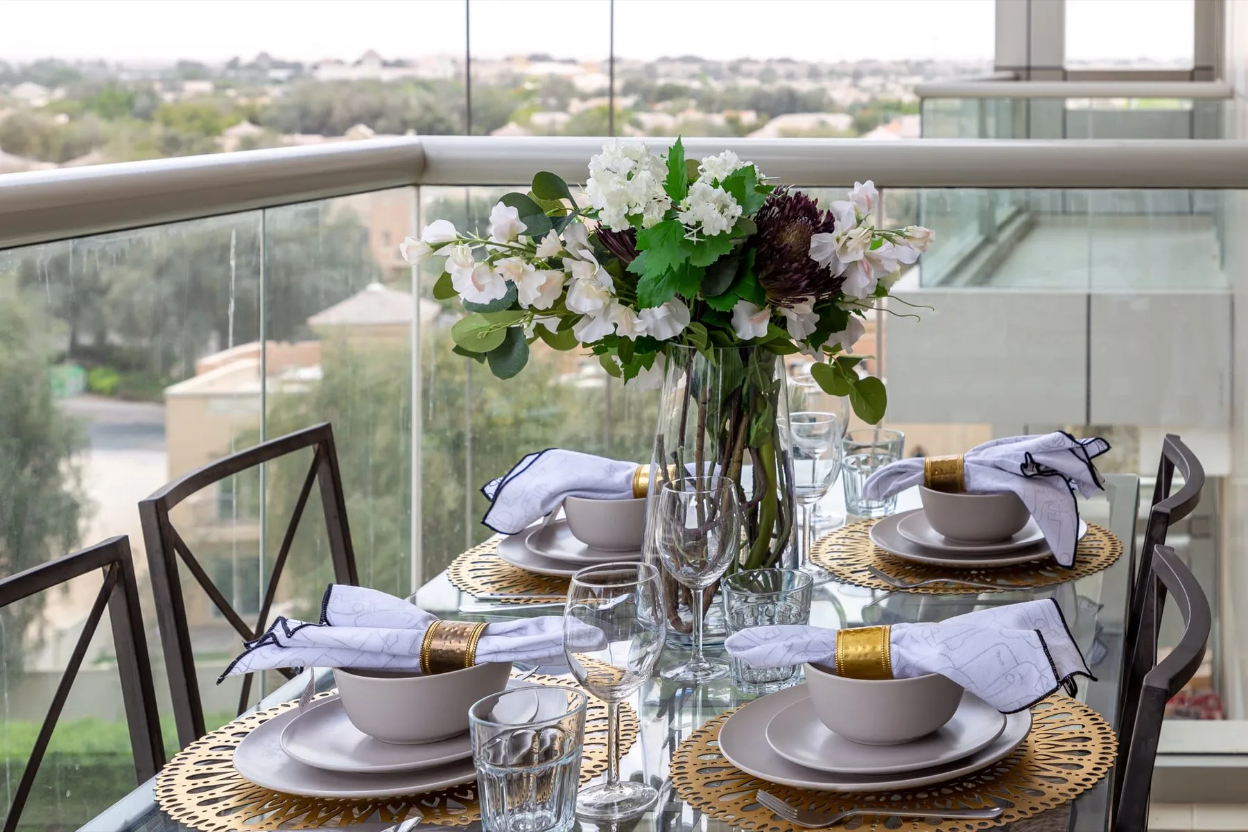 Una mesa de comedor está preparada con platos, cristalería y flores junto a grandes ventanales.