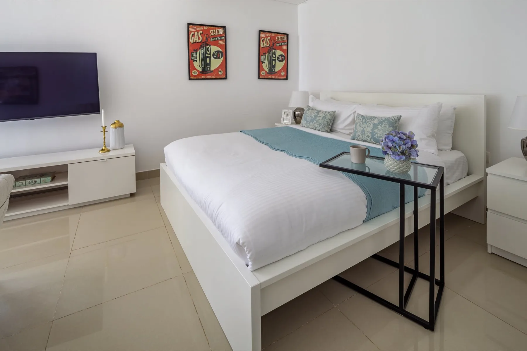 Un dormitorio luminoso con una cama blanca, mesas auxiliares y un televisor.