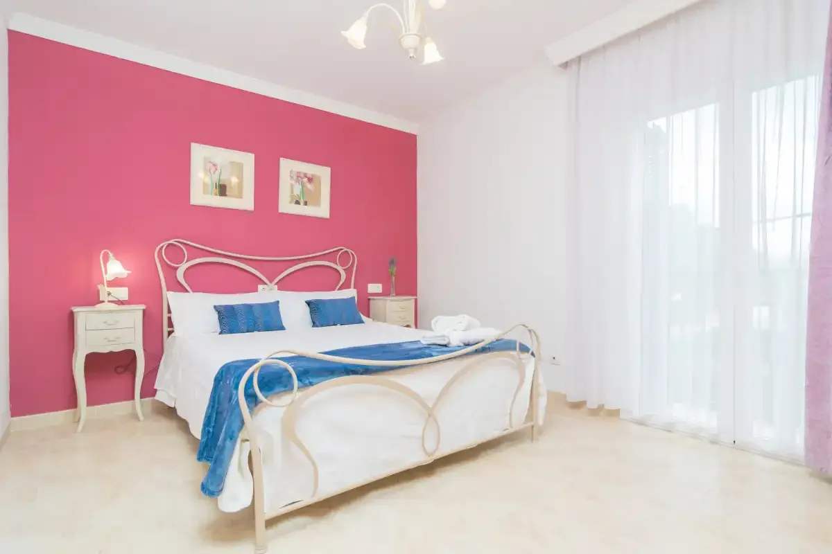 A bedroom with a white bed against a pink wall and sheer curtains.