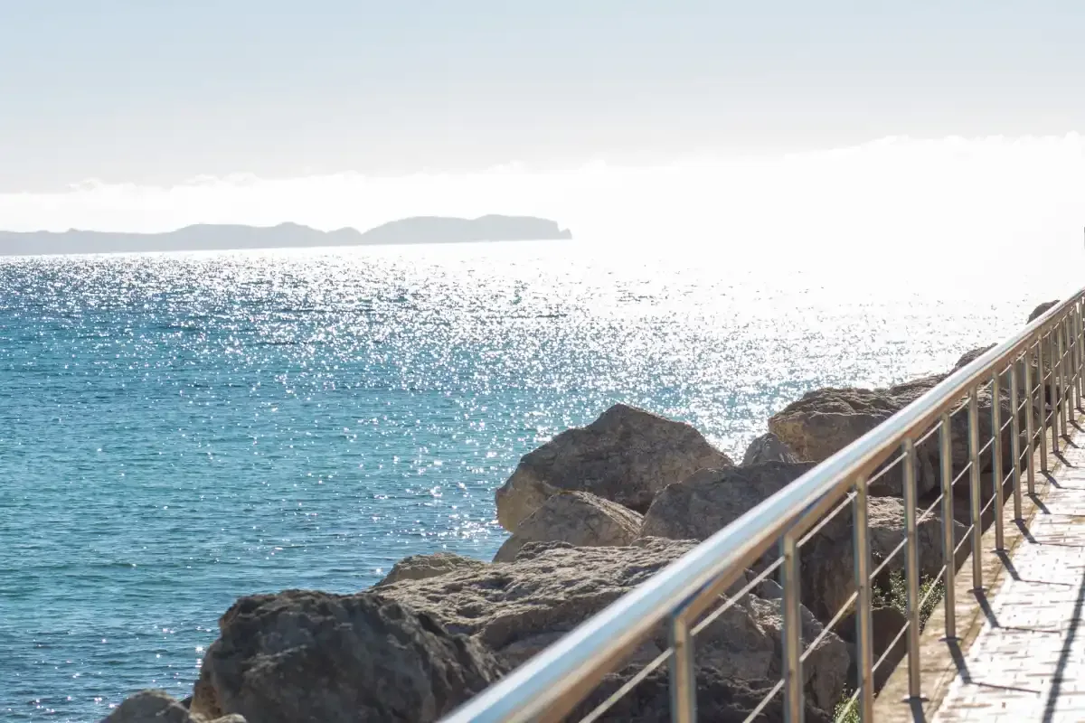 A seaside walkway with a metal railing runs beside rocky shoreline and bright blue water.