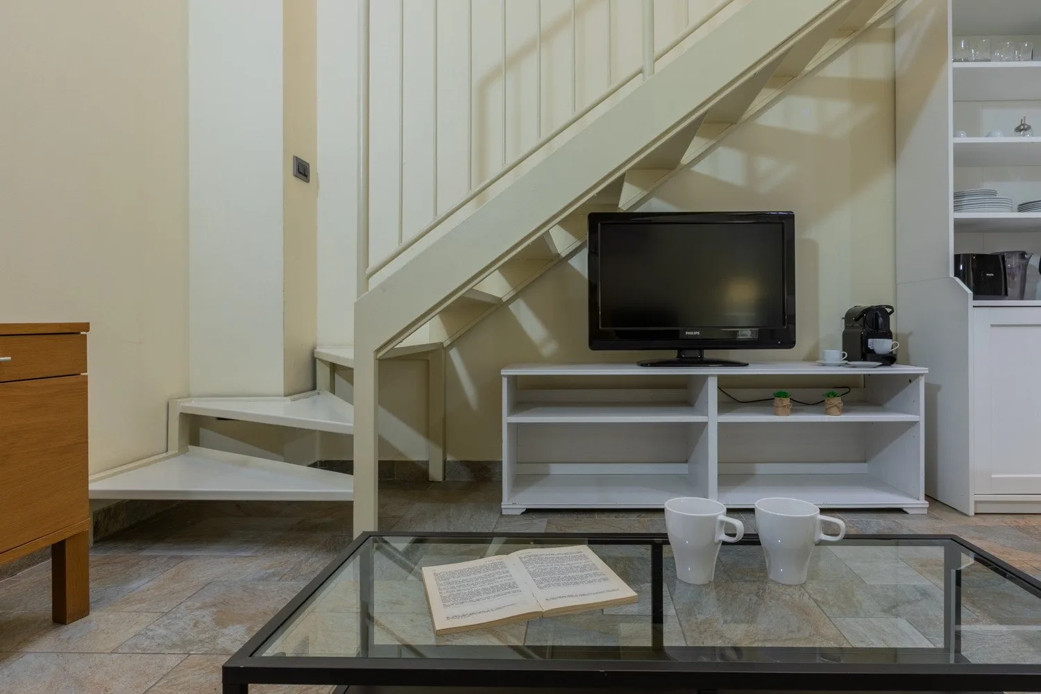 Una sala de estar con un televisor bajo una escalera y una mesa de centro de vidrio.