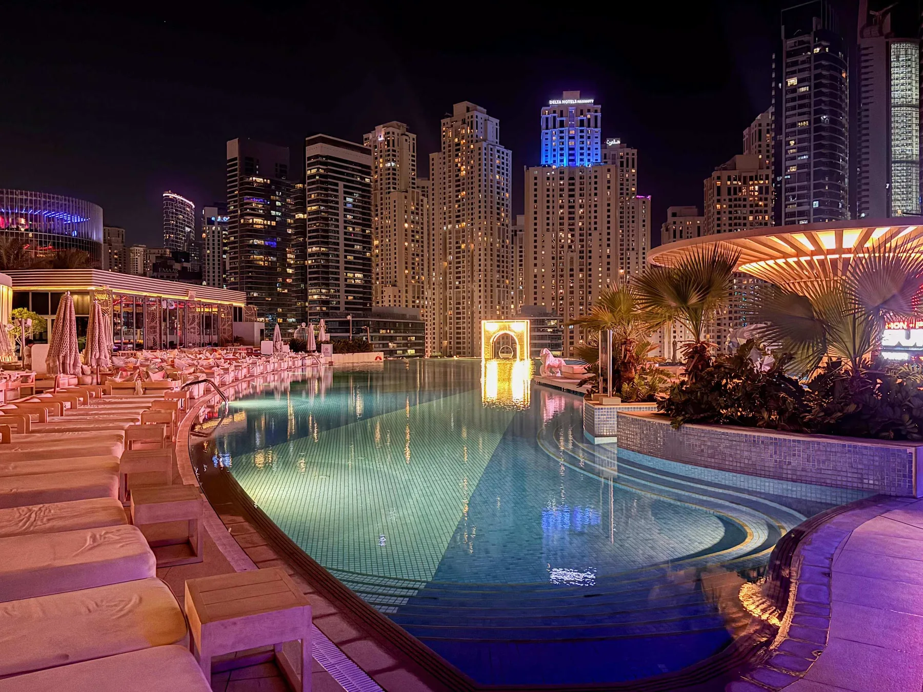Una piscina exterior iluminada está rodeada de asientos y edificios urbanos iluminados por la noche.