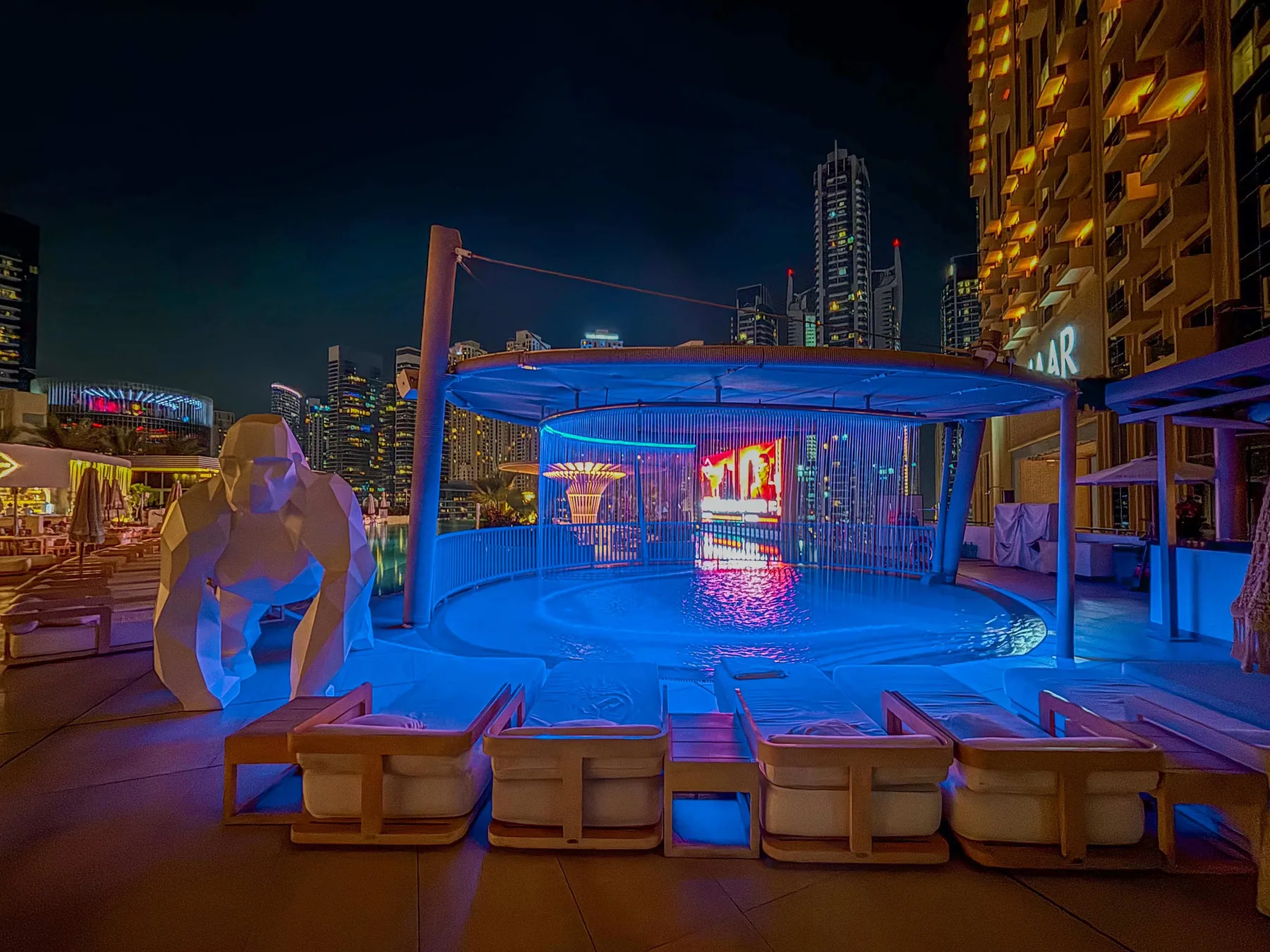 Una zona de piscina en una azotea brilla en azul por la noche, con asientos y edificios al fondo.