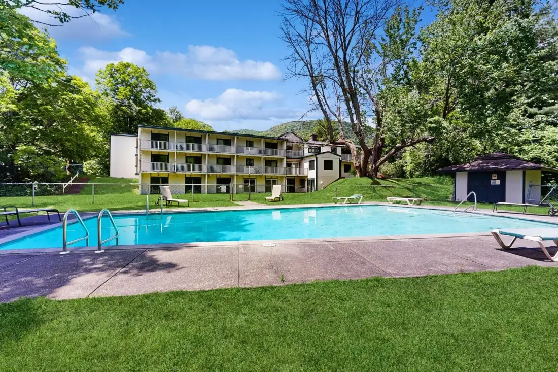 Una piscina al aire libre está junto a un edificio de varias plantas, rodeada de árboles.