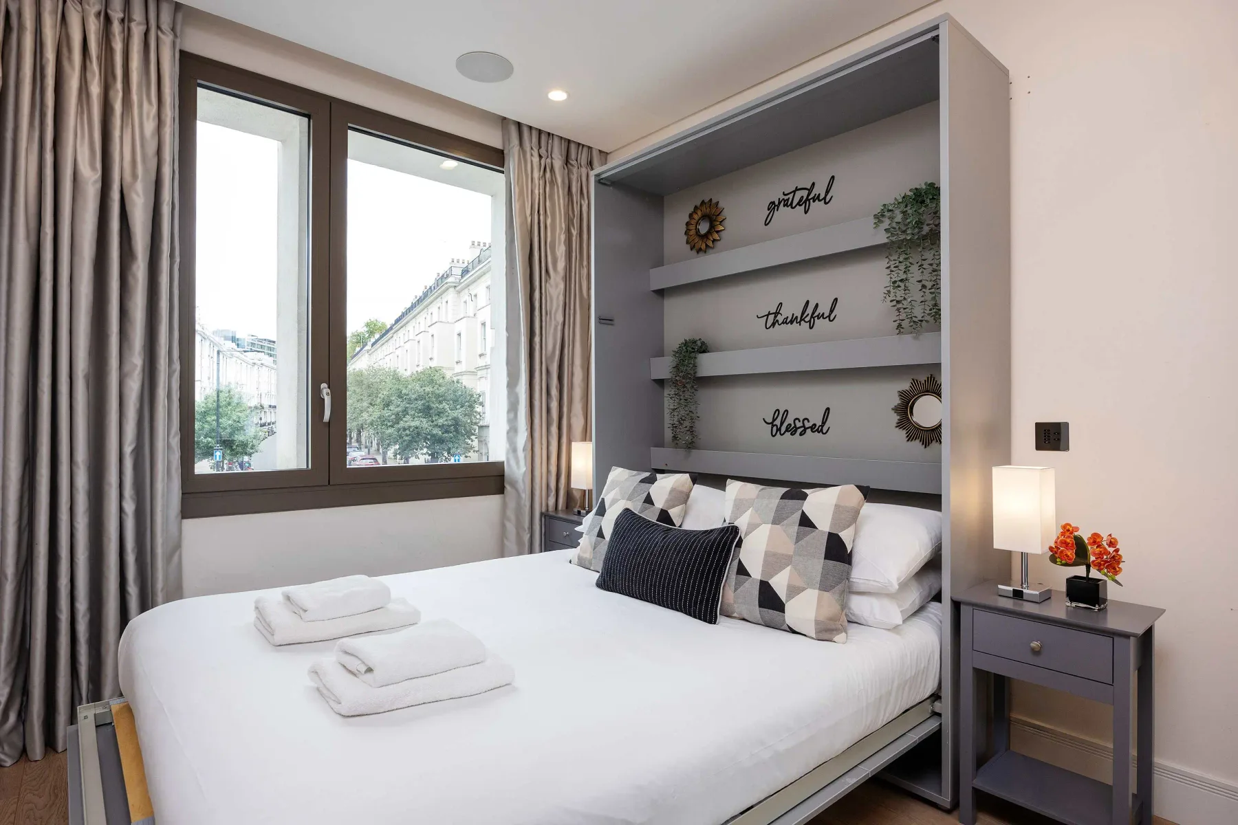 A bedroom with a bed beneath decorative wall shelves and beside a large window.