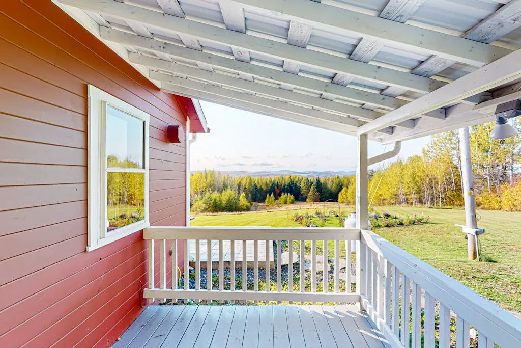 A covered porch with white railings overlooks a grassy yard and tree line.