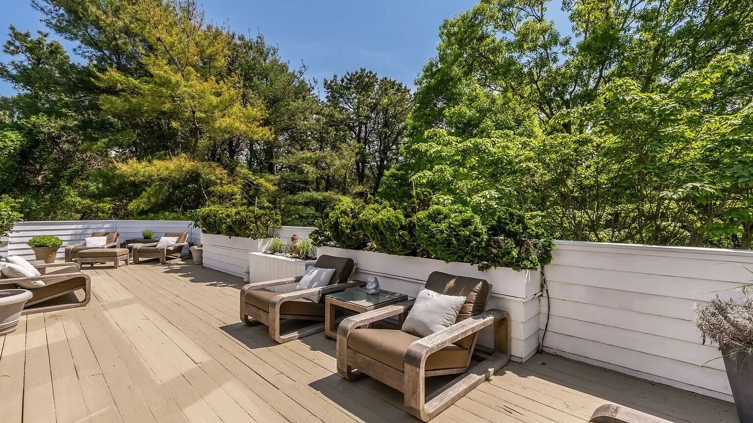 Una terraza exterior tiene asientos con cojines junto a un muro blanco con jardineras y árboles frondosos.