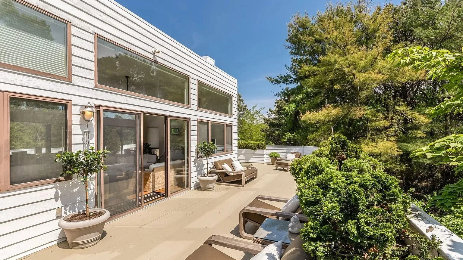 Un patio soleado junto a una casa moderna, con asientos y vegetación frondosa.