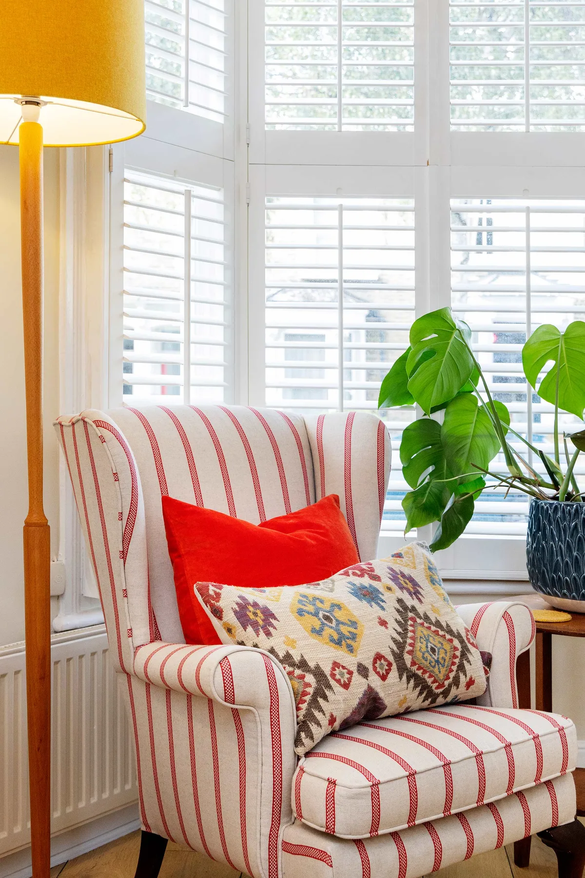 Un sillón a rayas con cojines coloridos está junto a una planta frondosa frente a ventanas luminosas.