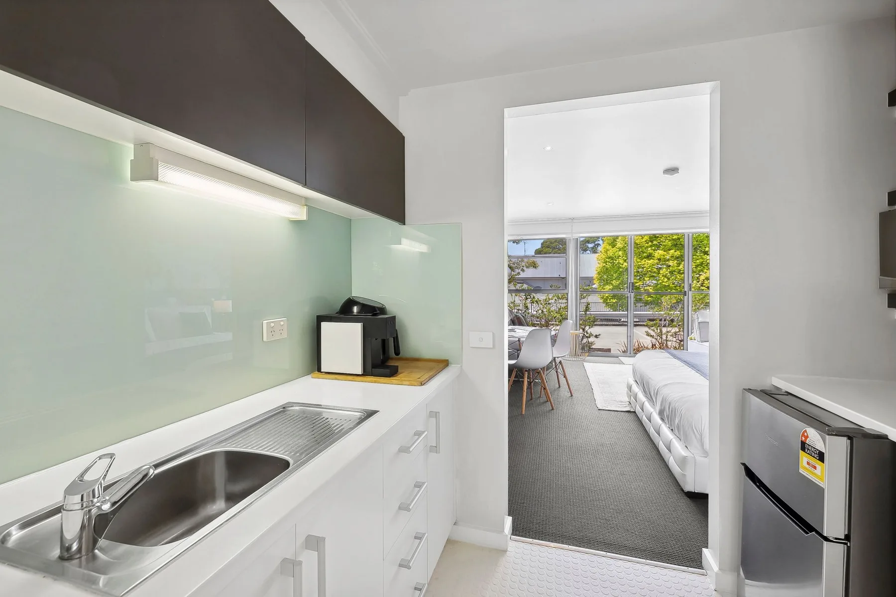 A compact kitchenette opens onto a bedroom with a bed and bright windows.