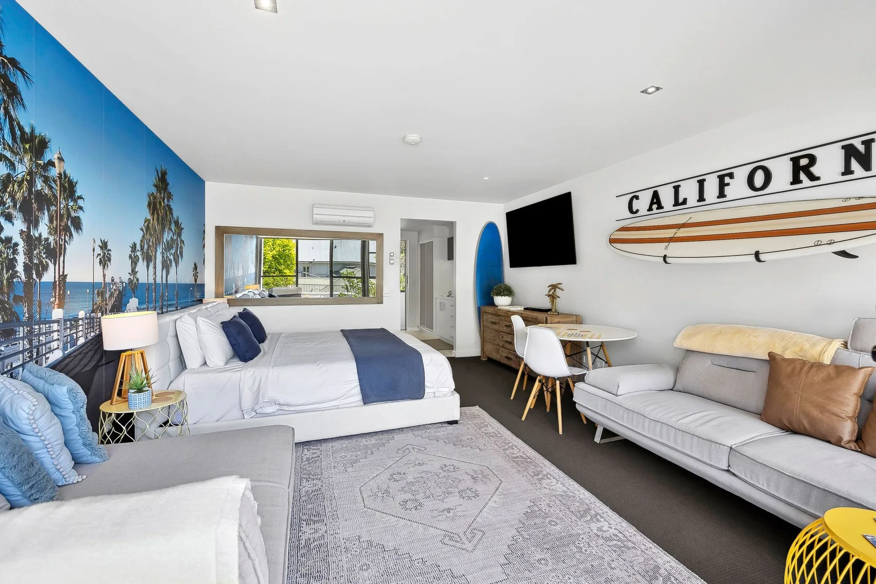 A bright motel room with a bed, seating, and a surfboard wall decoration.