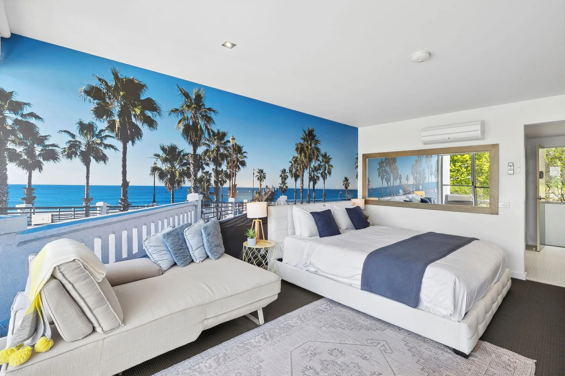 A bright bedroom with a bed, seating, and a large beach mural.
