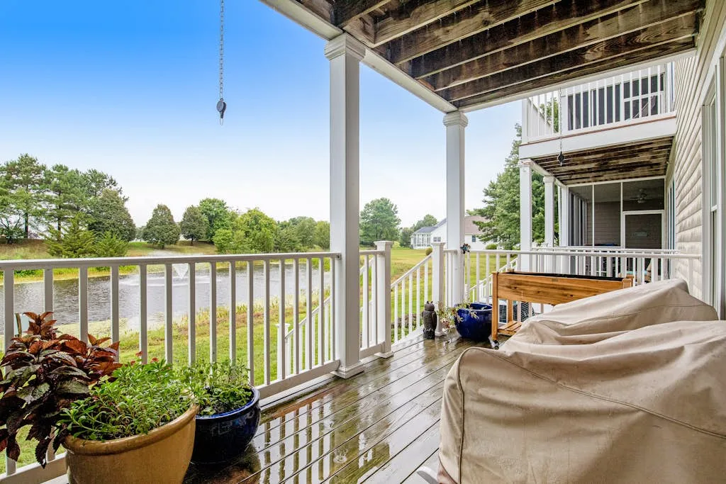 A covered outdoor deck with seating, potted plants, and a railing overlooking greenery.