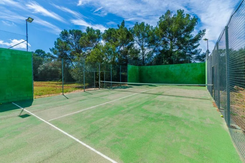 An outdoor tennis court with green walls and fencing beneath a blue sky.