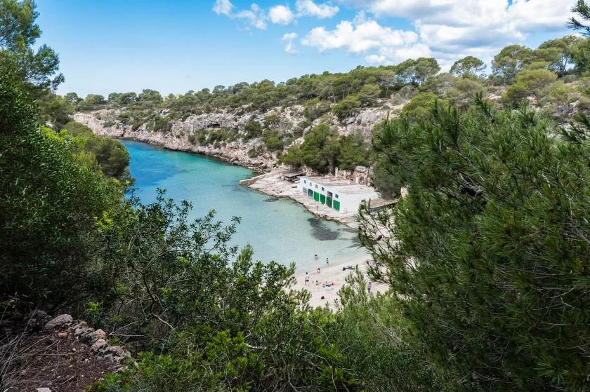 A turquoise cove with a small sandy beach and rocky, tree-covered shores.