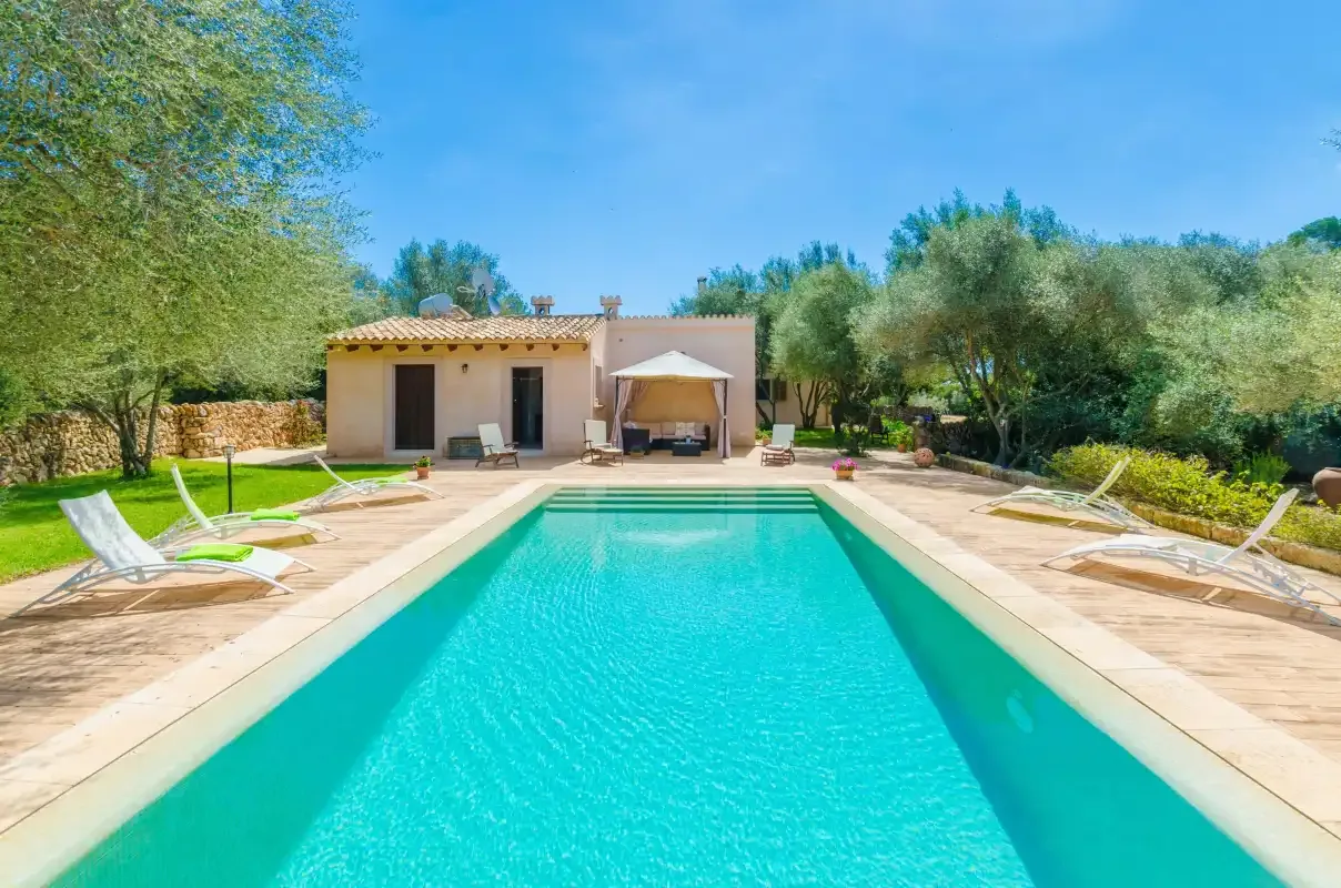 A swimming pool and surrounding patio sit in front of a low house.