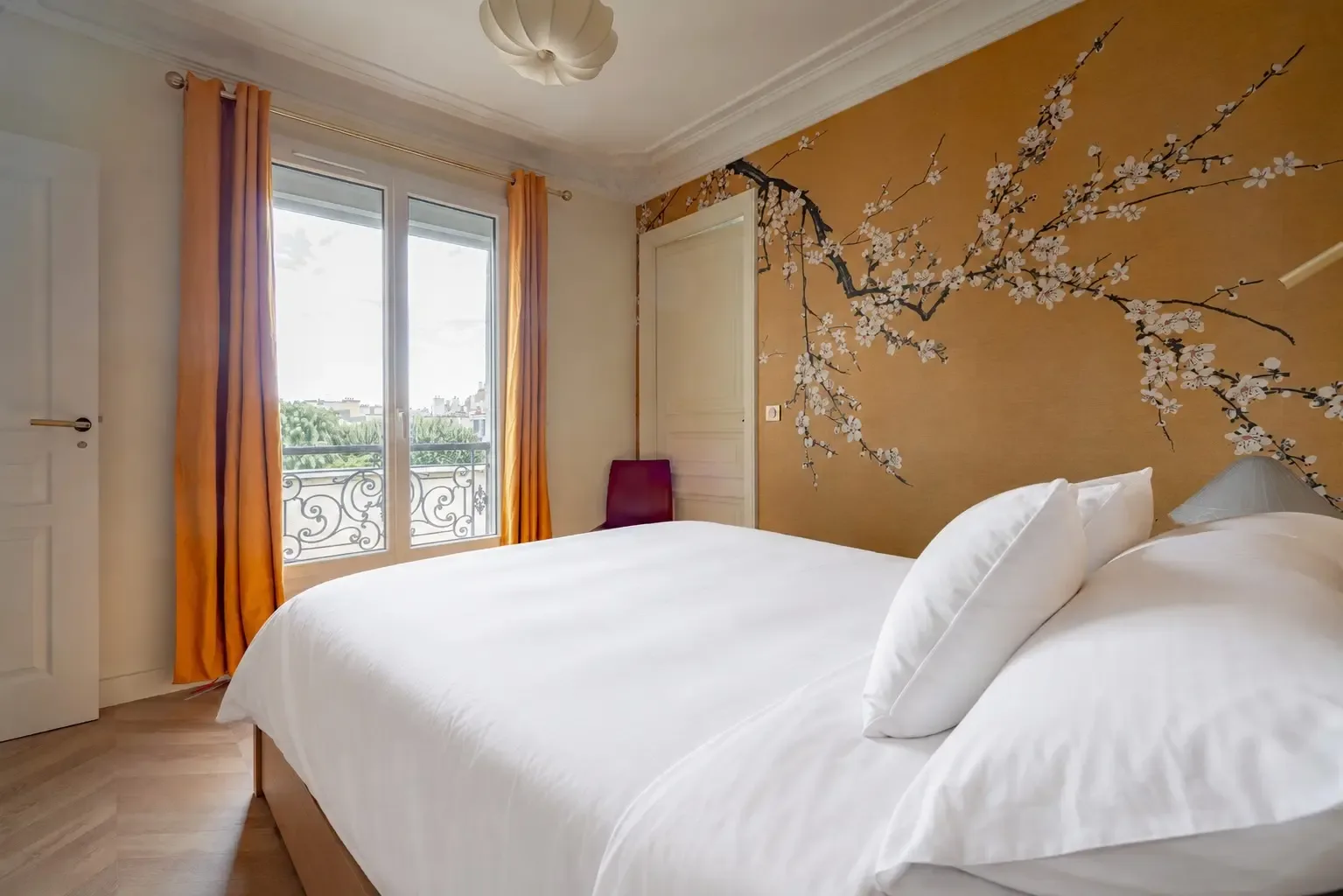 Un dormitorio con una cama blanca, cortinas naranjas y una rama florida decorativa en la pared.